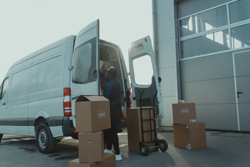 A courier loads cardboard parcels into the open rear doors of a white cargo van parked outside a depot loading bay, with a hand truck and stacked boxes beside him.