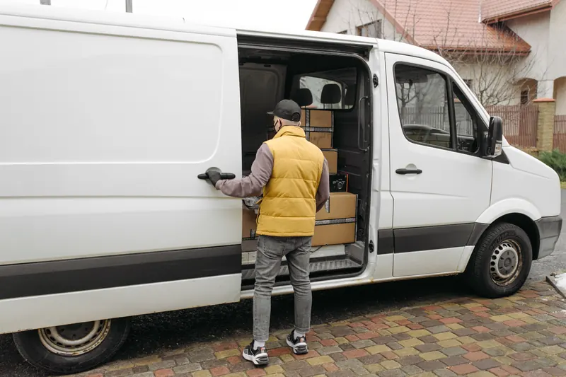 A delivery driver in a yellow vest slides open the side door of a white van stacked with parcels, parked on a paved residential driveway.