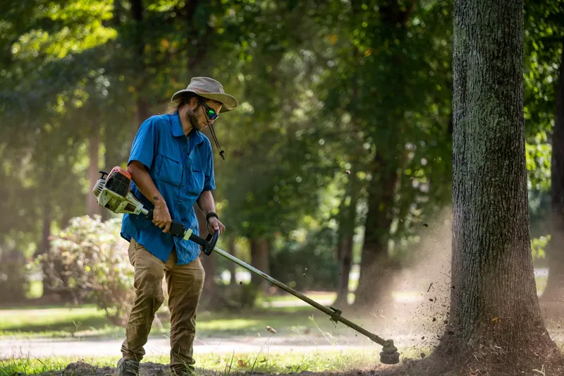A landscaper in a sun hat and safety glasses edges the grass around a tree trunk with a string trimmer, dust and clippings caught in the afternoon light.