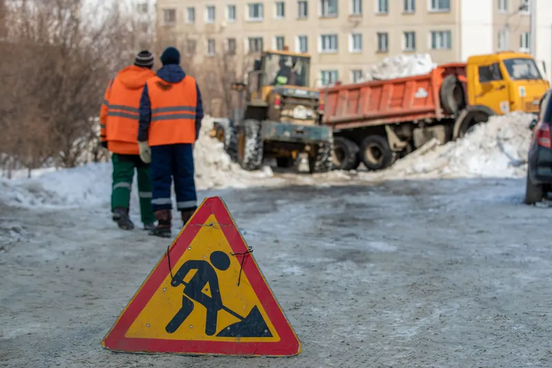 Two public-works crew members in orange high-visibility vests walk toward a wheel loader filling a tipper truck with cleared snow, a roadworks warning triangle in the foreground.