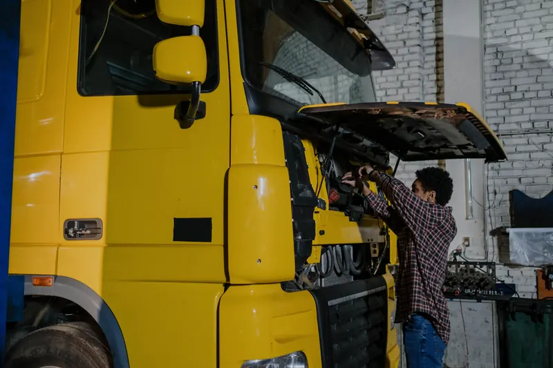 A mechanic in a plaid shirt reaches into the open front of a yellow heavy truck inside a whitewashed brick workshop.