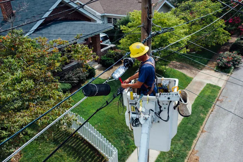 A lineworker in a yellow hard hat works from an aerial-lift bucket on an overhead cable splice above a tree-lined suburban street.