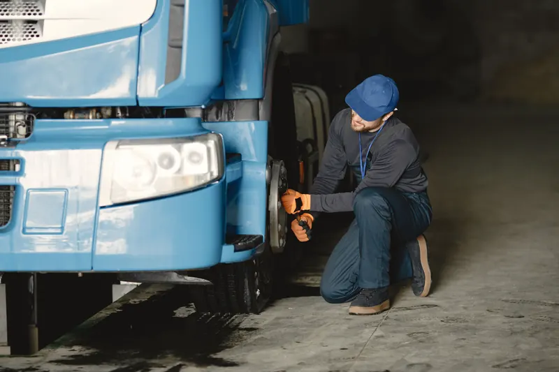 A technician in a blue cap and orange gloves kneels at the front wheel hub of a blue truck, working on the brake assembly in a dim fleet workshop.